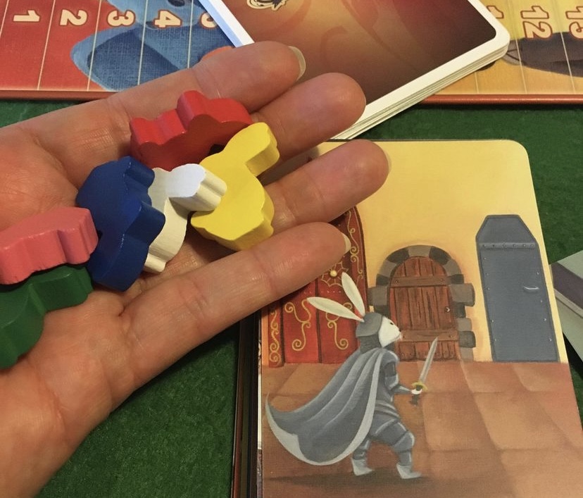 Image of a white hand holding six different coloured rabbit meeples with a card of a white rabbit in shining armour facing three different doors (from the tabletop game DIXIT)